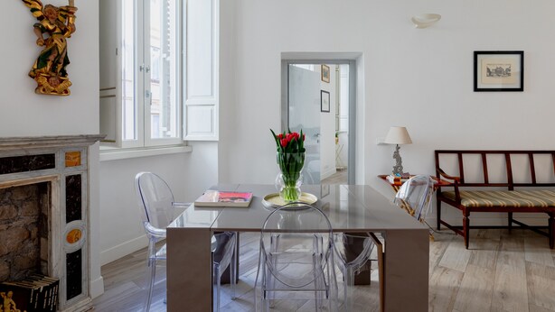 A modern dining area with a glass table, clear chairs, a vase of red flowers, and a fireplace with books.