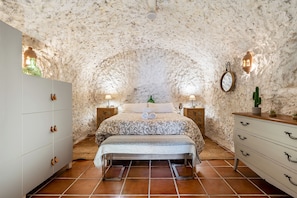A bedroom with a stone wall, a bed with a patterned bedspread, two bedside tables with lamps, a wooden dresser, and a bench.