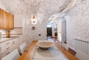 A rustic interior with a wooden table, white chairs, a woven bowl centerpiece, and a stone wall.