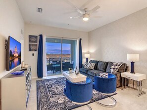 A modern living room with a flat-screen TV, a sofa, two blue chairs, a coffee table, and a balcony with a view.
