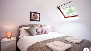 A bedroom with a bed, pillows, a bedside lamp, a framed picture, a window, and a towel on the bed.