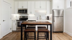 A modern kitchen with white cabinets, stainless steel appliances, and a wooden table with two stools. - Downtown Escape 1BR Condo (Traverse City)