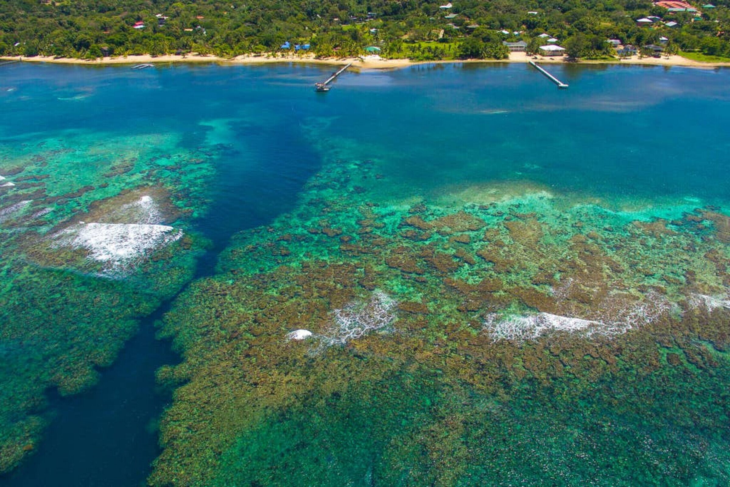 An aerial view of a coastal area with clear turquoise waters, visible coral reefs, and a small pier.