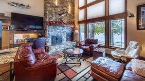 A cozy living room with a stone fireplace, leather sofas, a flat-screen TV, and large windows offering a view of the outdoors. - Lake House on Lake Blaine (Kalispell)
