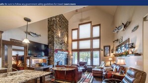 A spacious living area with a stone fireplace, large windows, and a television. - Lake House on Lake Blaine (Kalispell)