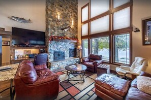 A cozy living room with a stone fireplace, leather sofas, a flat-screen TV, and large windows offering a view of the outdoors. - Lake House on Lake Blaine (Kalispell)