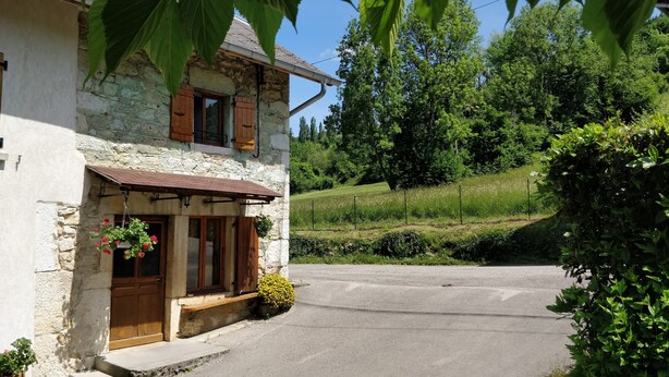 Une maison en pierre avec une porte et des fenêtres en bois, un petit porche orné de jardinières suspendues et une allée pavée menant à une zone herbeuse.