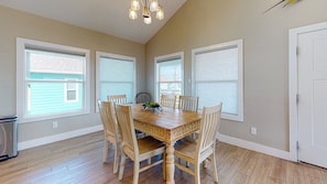 A dining room with a wooden table and six chairs, large windows with blinds, and a chandelier.
