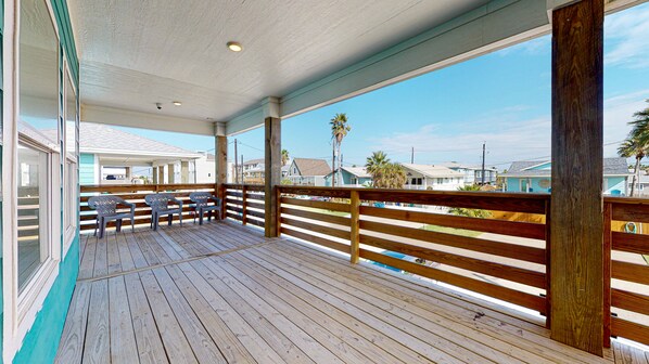A wooden deck with a railing, outdoor seating, and a view of houses and palm trees.