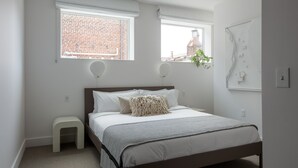 Ein Schlafzimmer mit einem Bett, einem kleinen Hocker und zwei Fenstern.