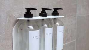 Trois flacons en verre transparent, dotés de pompes doseuses et d’étiquettes blanches, sont posés sur une étagère blanche, devant un mur carrelé.