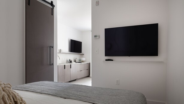 Une chambre à coucher avec un lit, un téléviseur fixé au mur, une porte coulissante et un coin cuisine à l’arrière-plan.