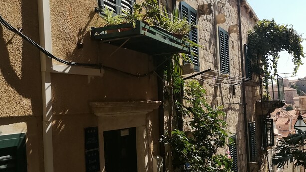 Una calle estrecha con edificios de fachadas de piedra y contraventanas verdes. Hay macetas con plantas en los balcones y una pequeña entrada con una puerta.