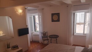 Un dormitorio con una cama, un televisor, una silla, una mesita y una ventana con cortinas.