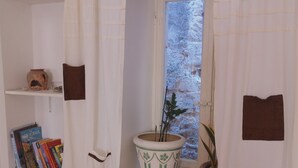 Un baño con paredes blancas, una ventana con vidrio esmerilado, una estantería con libros y una planta en maceta, y una cortina de ducha con detalles marrones.