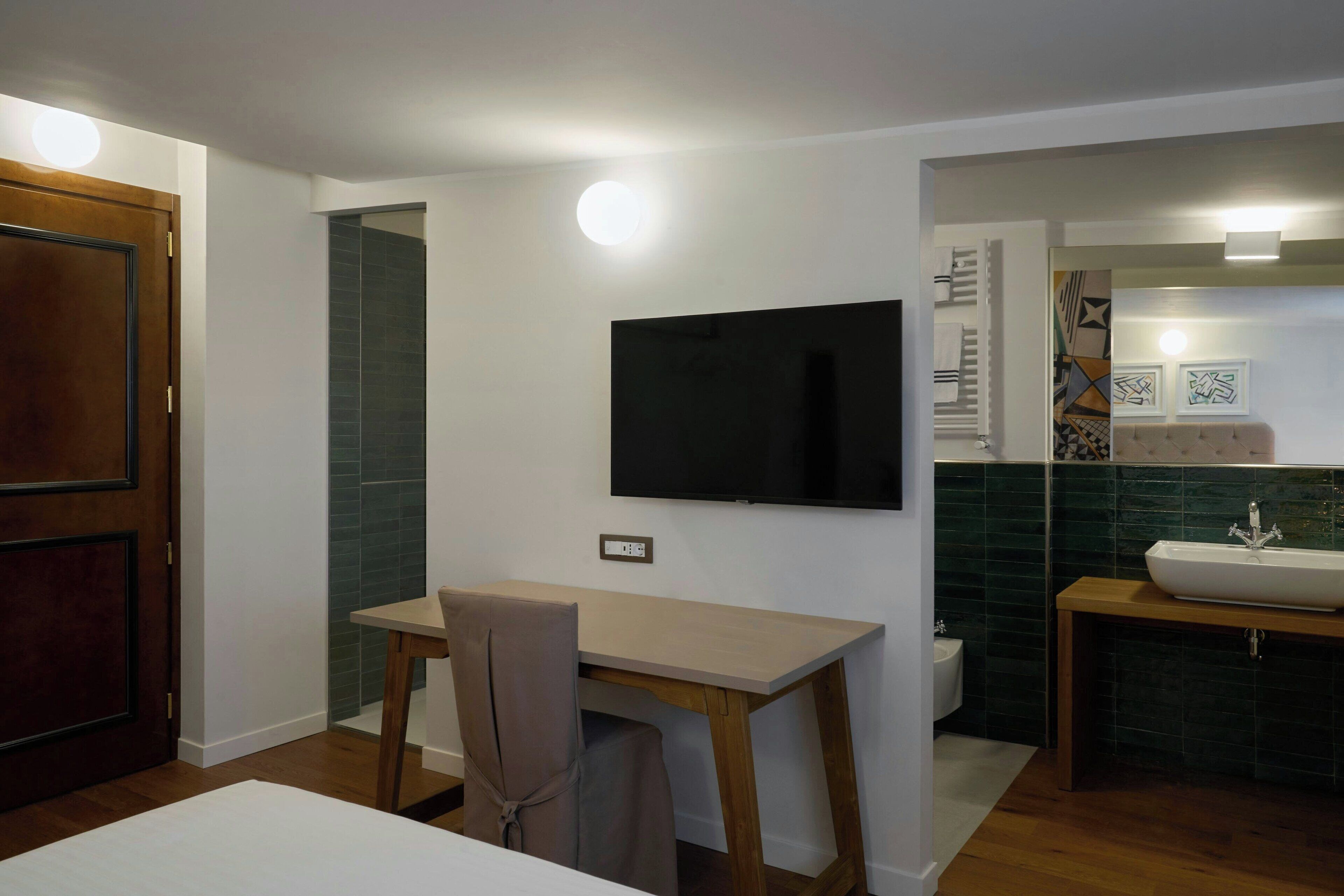 A compact room with a wooden desk, a chair, a television mounted on the wall, a sink, and a toilet.