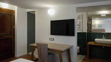 A compact room with a wooden desk, a chair, a television mounted on the wall, a sink, and a toilet.