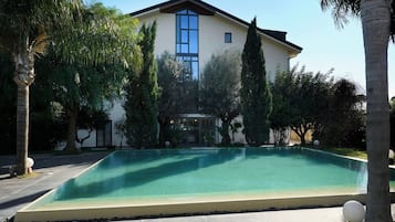 A rectangular swimming pool with a clear blue water surface, surrounded by a paved deck. In the background, there is a two-story building with a beige facade and a black roof.