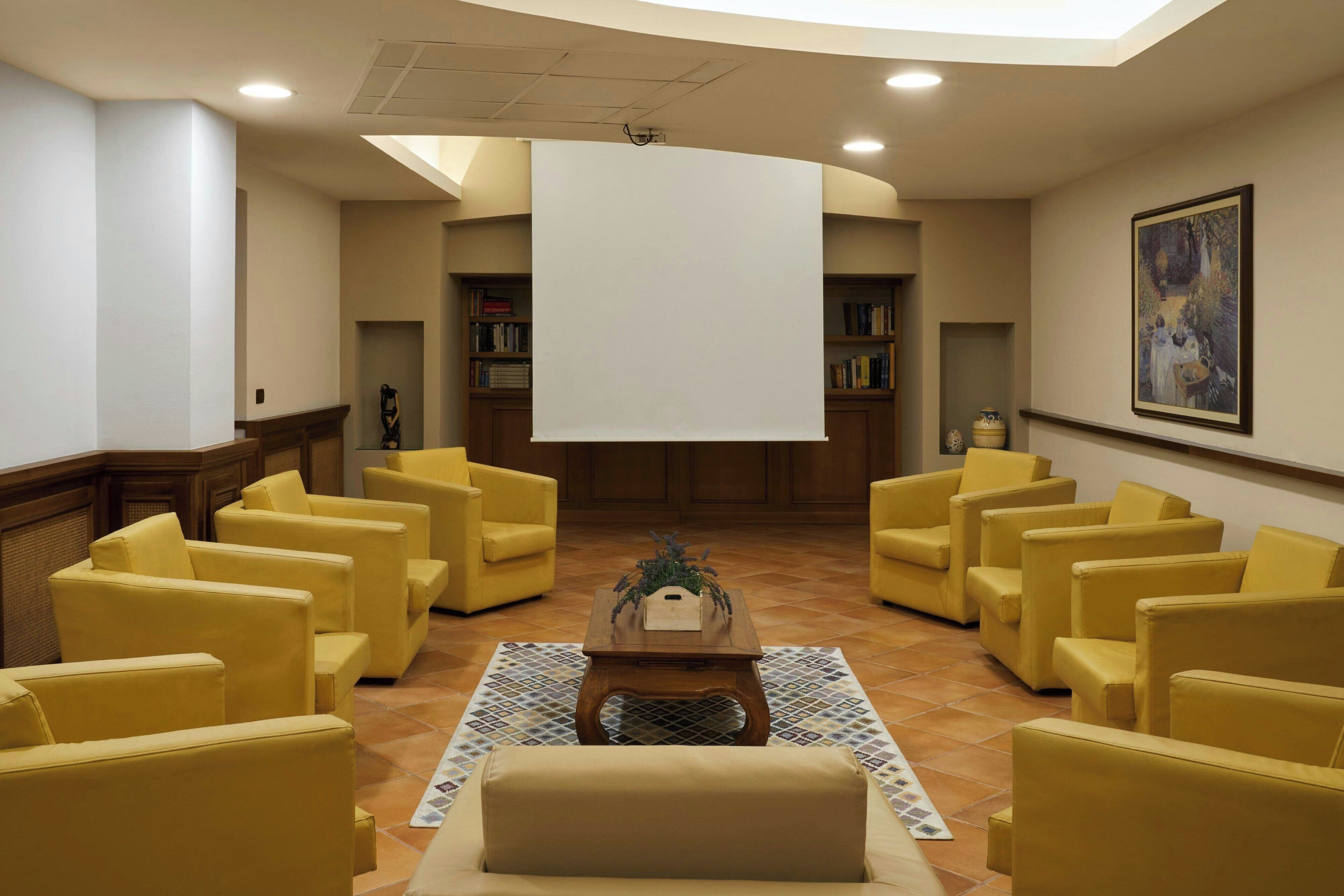 Un vestíbulo con sillones amarillos dispuestos en semicírculo, una mesa de centro y una pantalla blanca grande.