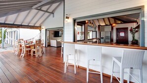 Een ruim overdekt terras met houten vloeren, witte rieten barstoelen en een lange eettafel met stoelen.