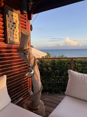 A wooden deck with a rustic sculpture, white cushions, and a view of the ocean.