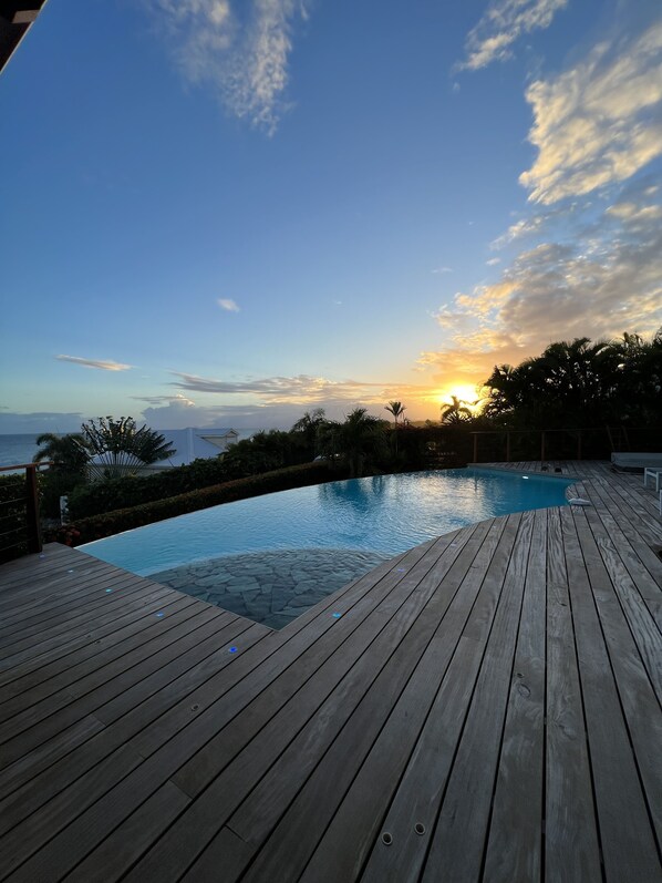 A wooden deck with a swimming pool, surrounded by greenery and overlooking a body of water.