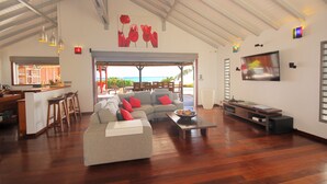 Een ruime woonkamer met een bank, salontafel en televisie. Er is een keuken met een bar en krukken. De kamer heeft een hoge plafond met zichtbare balken en een groot raam met uitzicht op de oceaan.