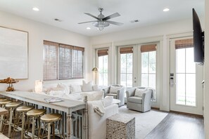 A spacious living room with a high ceiling fan, large windows with blinds, a bar area with stools, and a television mounted on the wall.