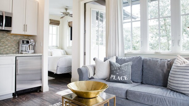 A modern kitchen with a coffee maker, a dining area with a sofa and pillows, and a view of the outdoors through large windows. - Carriage House | Walk to Dining and Beach (Panama City Beach)