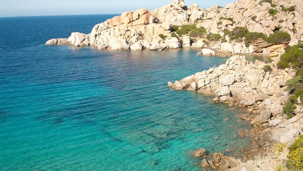 A coastal landscape with clear turquoise waters, rocky cliffs, and sparse vegetation.