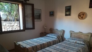 A bedroom with two wooden beds, a small bedside table, a lamp, and a window with a view of trees and a stone wall.