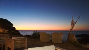 A wooden bench with a white cushion, wicker chairs, and a flagpole with a flag on a beach at sunset.