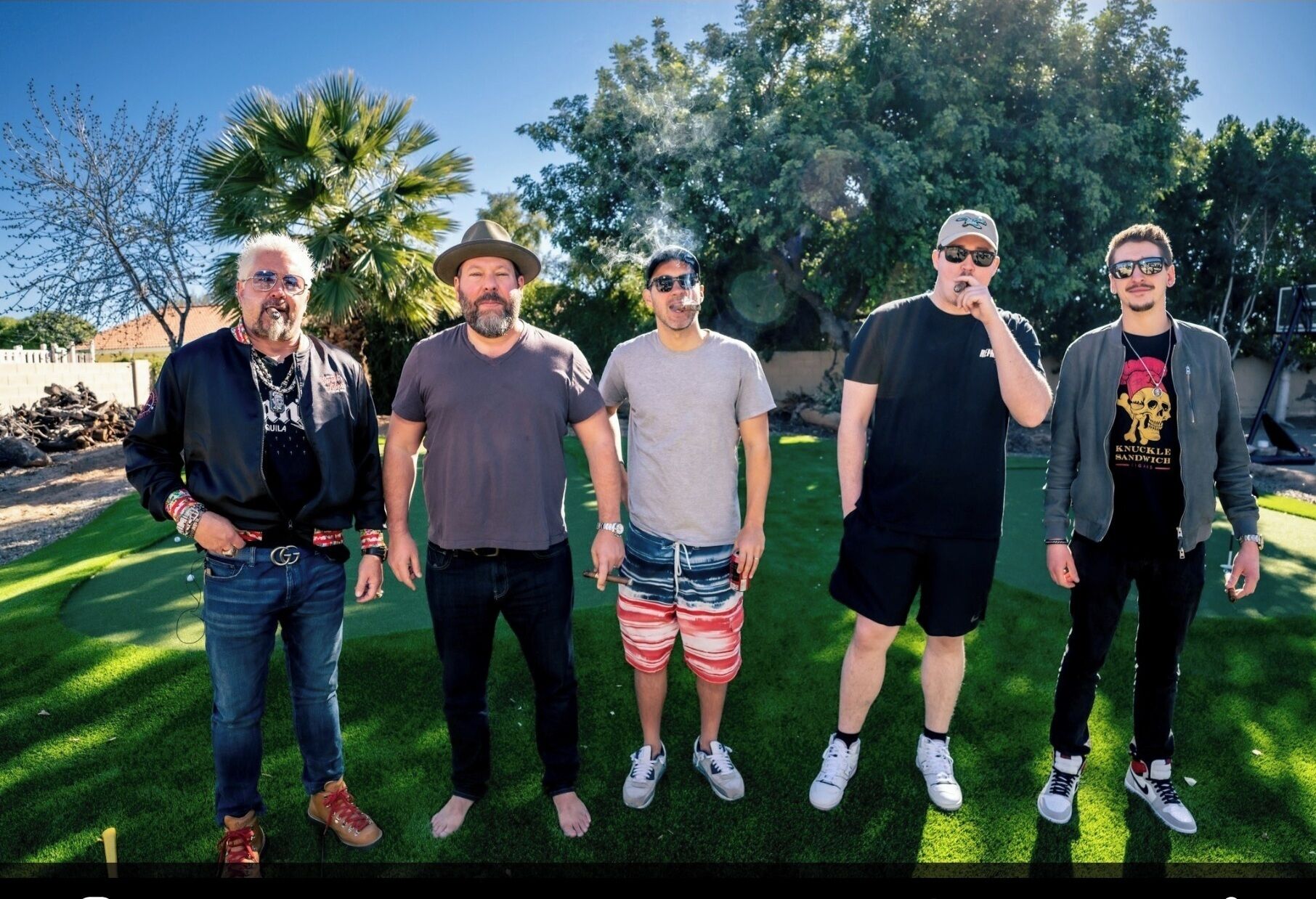 A group of five people standing outdoors on a grassy area with trees and a clear sky in the background.