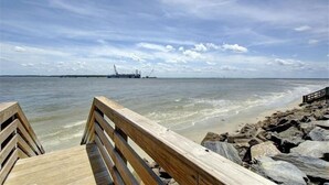 A wooden pier leading to a rocky beach with a view of the ocean and distant industrial structures. - Oyster Shell Cottage (St Simons Island)