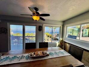 En matplats med ett bord, stolar, en takfläkt och stora fönster med utsikt över havet och palmerna.
