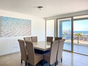 Uma sala de jantar com uma mesa grande e seis cadeiras, uma câmera de segurança instalada no teto e uma janela ampla com vista para a praia e o oceano.
