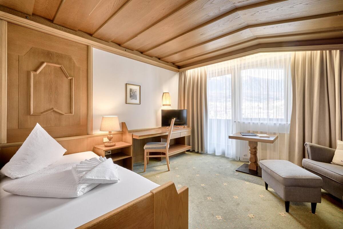 Quarto de hotel com teto de madeira, cama com roupa branca, escrivaninha com cadeira, televisão e janela com cortinas.