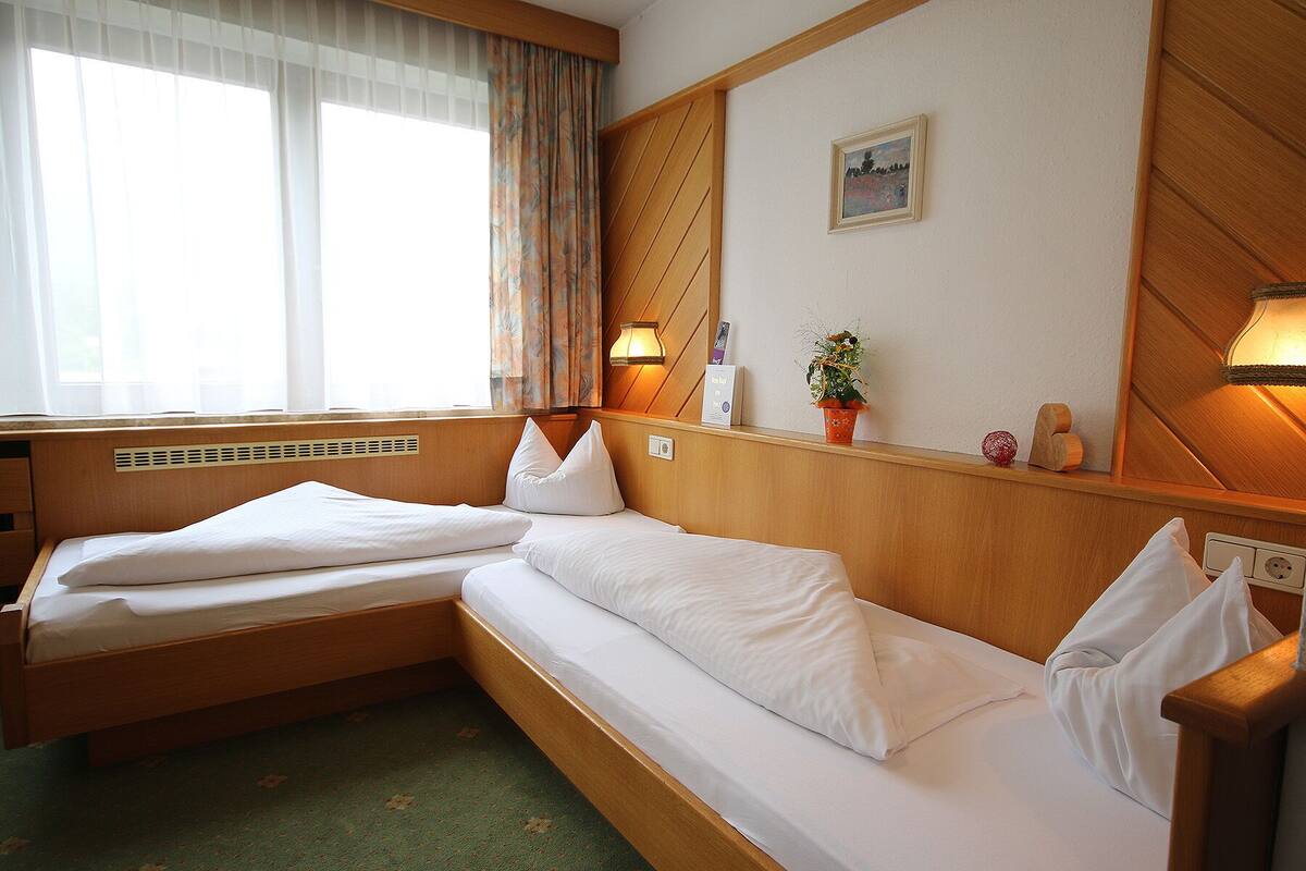 Quarto de hotel com duas camas de solteiro, uma janela com cortinas translúcidas, luminária presa à parede, pequena moldura com imagem, planta em vaso e cabeceira de madeira.