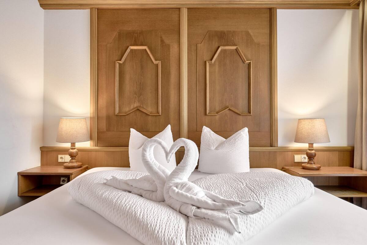 Quarto de hotel com uma cama grande, duas mesas de cabeceira com luminárias e um painel de cabeceira de madeira com arranjo de toalhas em formato de coração.