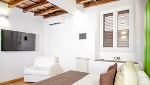 Een slaapkamer met een bed, een witte stoel, een televisie, een raam met zonwering en een radiator.