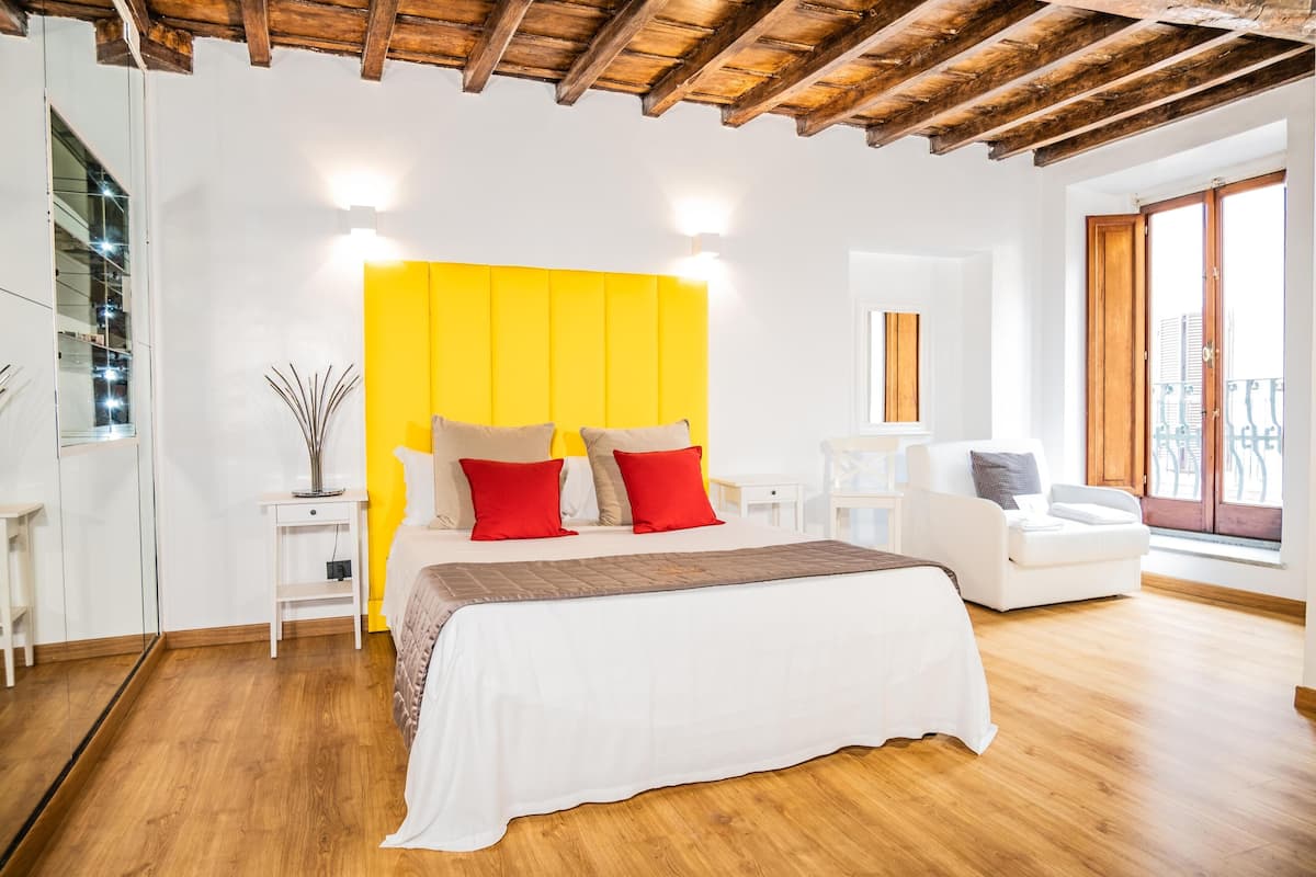 Quarto moderno com teto de madeira, cama com roupa de cama branca e almofadas vermelhas, sofá branco e uma janela grande com vista.
