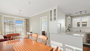 A dining area with a wooden table and chairs, a kitchen with white cabinets, and a large window with blinds.