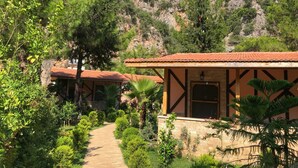 Un chemin menant à un bâtiment doté d’un toit en tuiles, entouré d’une végétation luxuriante et ayant une chaîne de montagnes en arrière-plan.