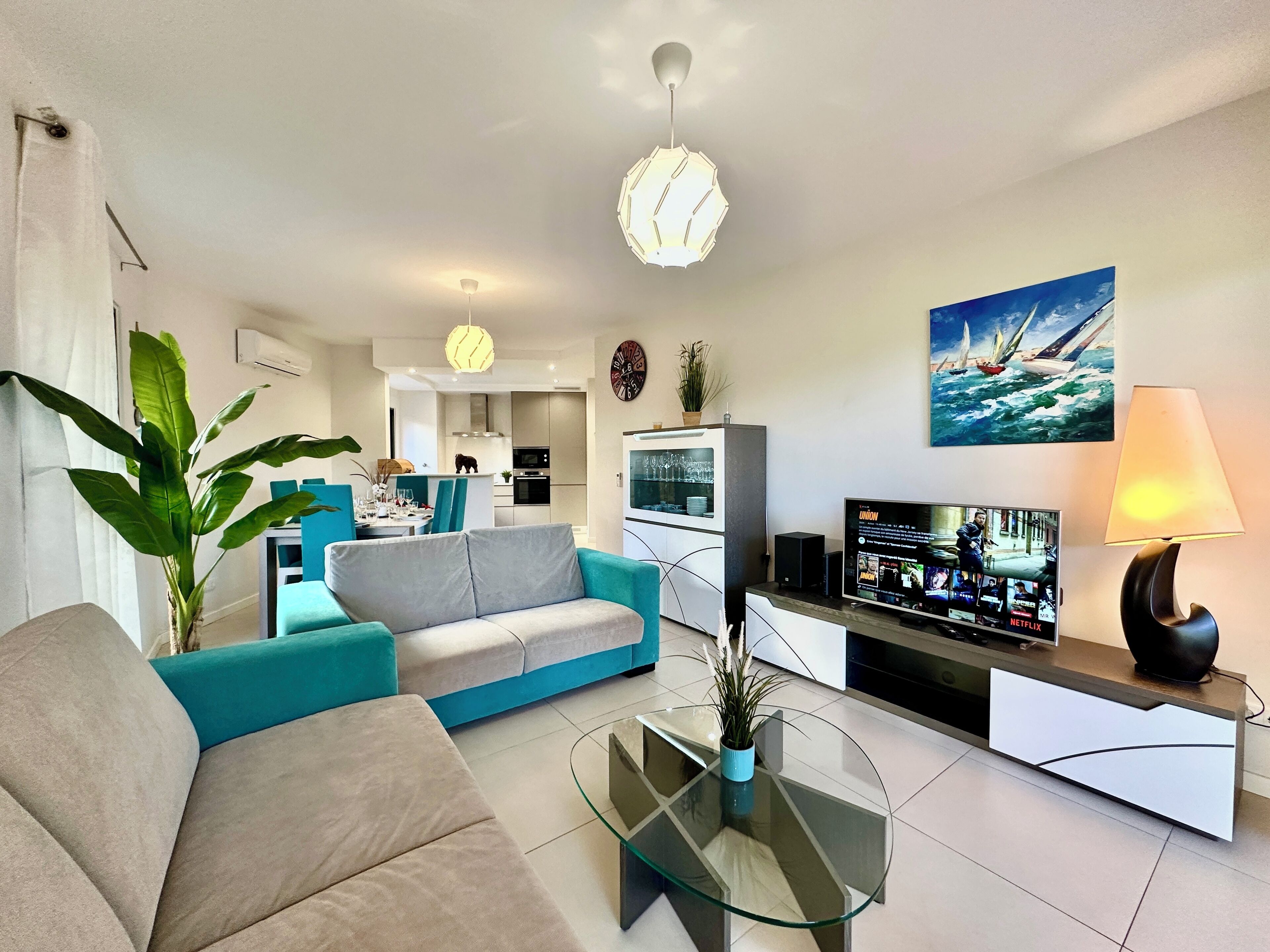 Ein modernes Wohnzimmer mit einer Couch, einem Glastisch, einem Fernseher und einer Küche im Hintergrund.