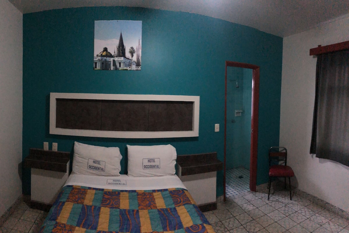 Una habitación de hotel con pared color esmeralda, una cama grande con colcha de colores, dos almohadas con la etiqueta "HOTEL OCCIDENTAL", un cuadro enmarcado de un edificio y una puerta que conduce a otra habitación.