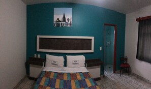 Une chambre d’hôtel avec un mur de couleur turquoise, un grand lit recouvert d’une couette aux couleurs vives, deux oreillers portant l’inscription « HÔTEL OCCIDENTAL », un tableau encadré représentant un bâtiment, et une porte donnant sur une autre chambre.
