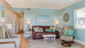 A living room with a brown leather sofa, a white coffee table, and a rocking chair. There is a clock on the wall and a painting above the sofa. - Oceanfront | Spacious Deck | Beautiful Views | Seagate Condo (Fernandina Beach)