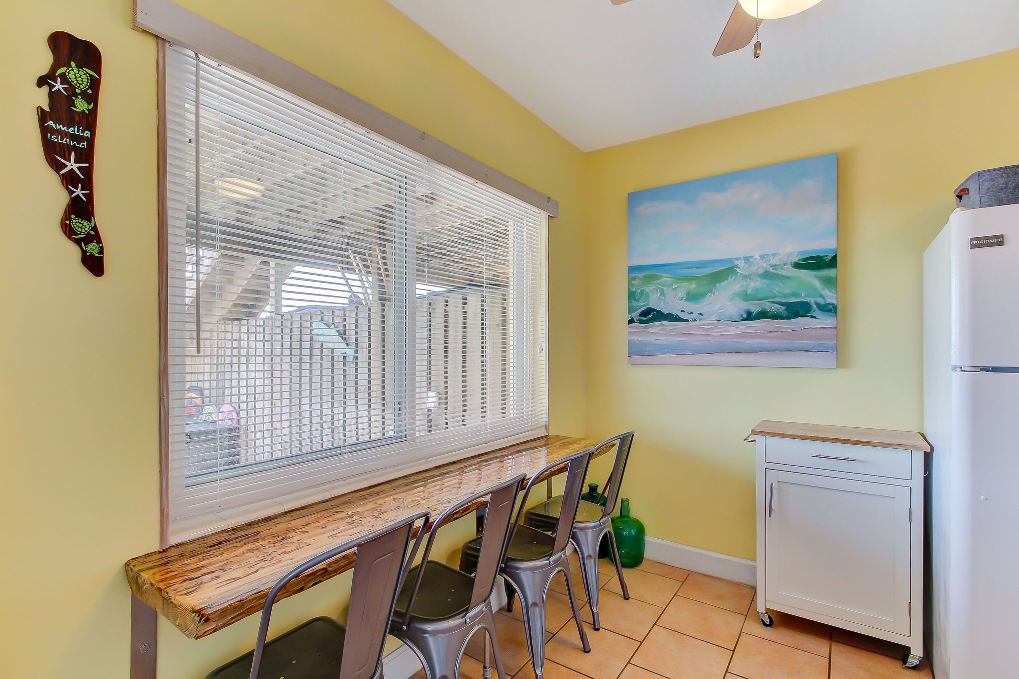 A dining area with a wooden bench and metal chairs, a window with blinds, a painting of waves, and a white cabinet.