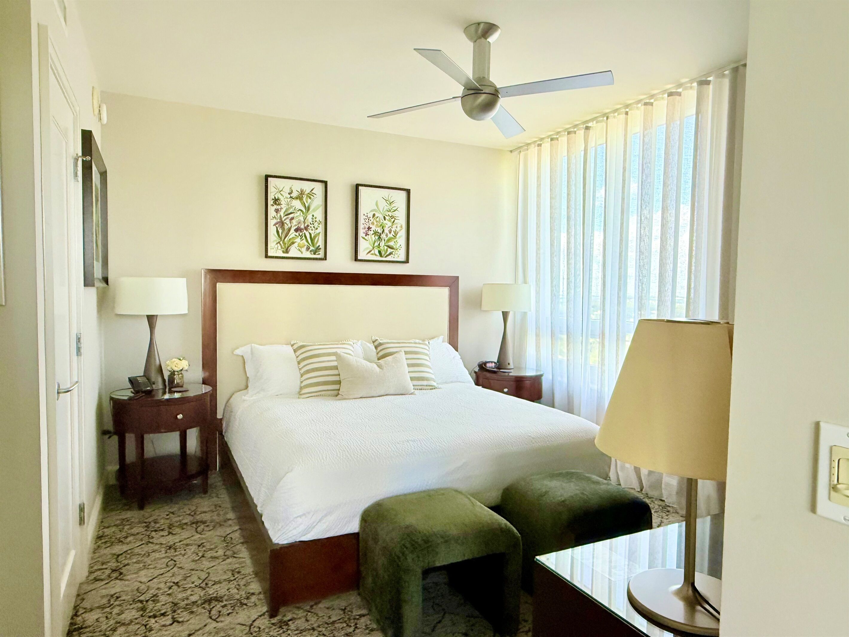 Une chambre d’hôtel avec un grand lit, deux tables de chevet avec des lampes, un ventilateur de plafond et une fenêtre avec des rideaux.