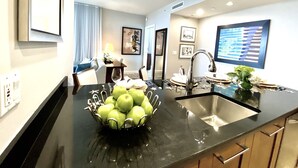 Eine moderne Küche mit schwarzem Arbeitsplatz, Spüle aus Edelstahl und einer Schüssel grüner Äpfel. An der Wand befinden sich gerahmte Bilder und im Hintergrund ein Essbereich.